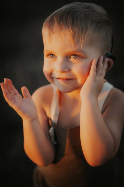 Gramm, a child with bilateral hearing loss, smiling for the camera and holding his cochlear implant sound processor. 