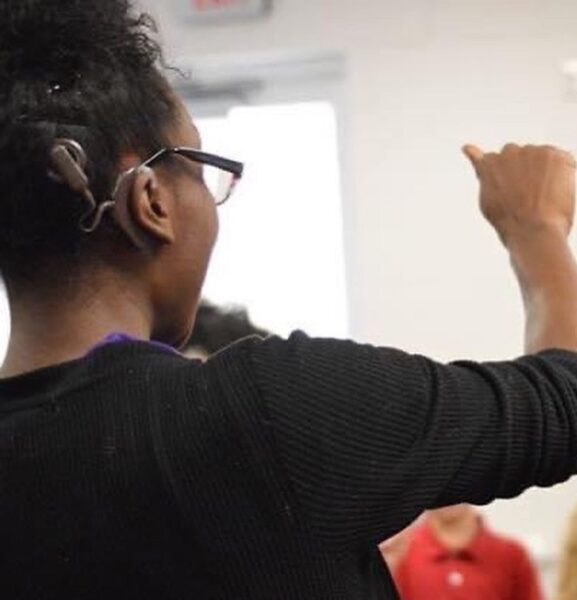 A picture of Kei-Che, a Hybrid Hearing Solution recipient, from behind showing her sound processor and her signing in ASL. 