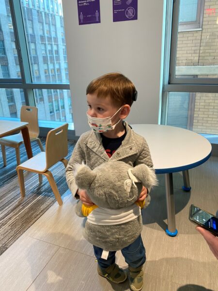 Theo who is a toddler with hearing loss, in the hospital with a Kaci the Koala stuffed animal and his cochlear implant. 