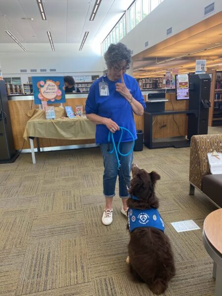 Donna Sue, who uses the Baha System for SSD, working a volunteer event at her local library and giving a command to her therapy dog. 