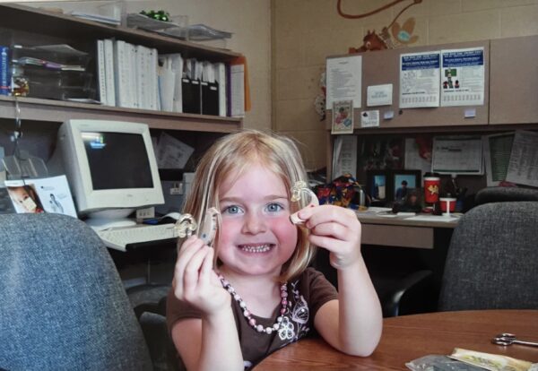 Morgan, who is a deaf young woman with cochlear implants, pictured as a child holding up two cochlear implant devices and smiling. 