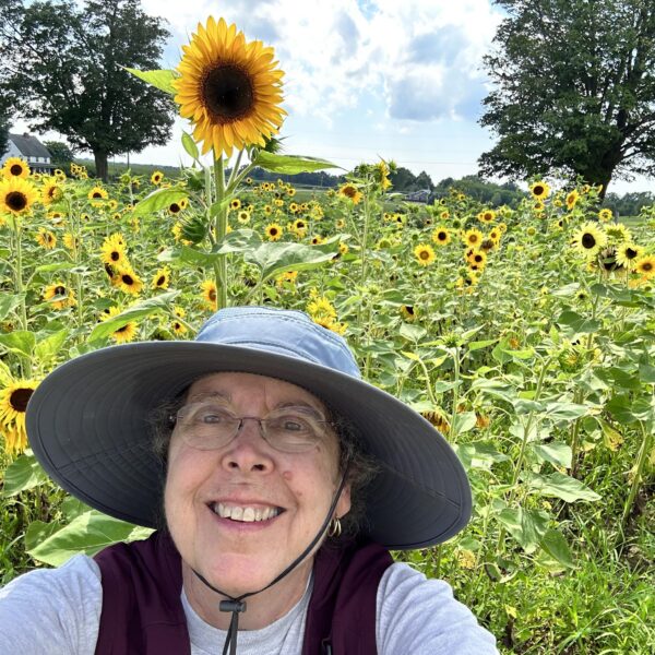 Margo stands in a field of sunflowers while wearing a sun hat with her cochlear hearing device.