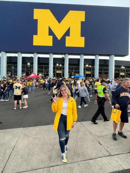 Morgan, a deaf young woman who has cochlear implants, posing in front of Michigan Stadium, holding "I Love You" in sign language. 