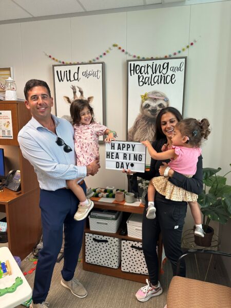 Matt & Jessica, who noticed signs of hearing loss in their children Sophie & Leah, posing for a family photo at the clinic, showing a sign that reads "Happy Hearing Day."