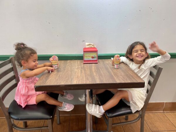 Sophie & Leah whose parents started noticing signs of hearing loss, posing for a picture sitting at a restaurant table, eating ice cream cones. 