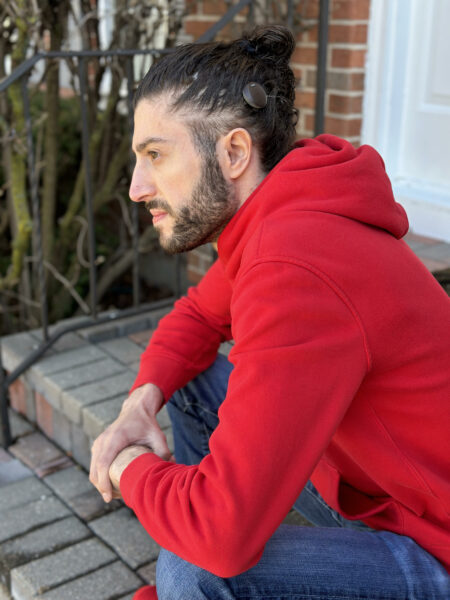 Peter, who has the Osia Sound Processor posing for a picture while sitting on a porch, wearing a red hoody. His Osia Sound Processor is visible in his hair. 