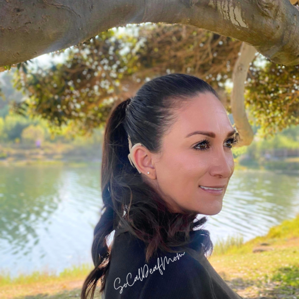 Sandra, who overcame her hearing loss with a cochlear implant, smiling for a side profile photo under a tree, standing next to a pond. 