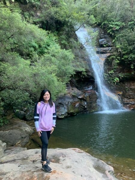 Nomiki, standing and smiling next to a waterfall; manage listening fatigue