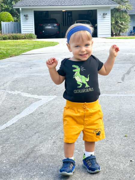 Jamie, a child who received cochlear implants to support his hearing health, smiling for a photo in a driveway with his hands up in the air and wearing a "cochlearsaurus" t-shirt. 