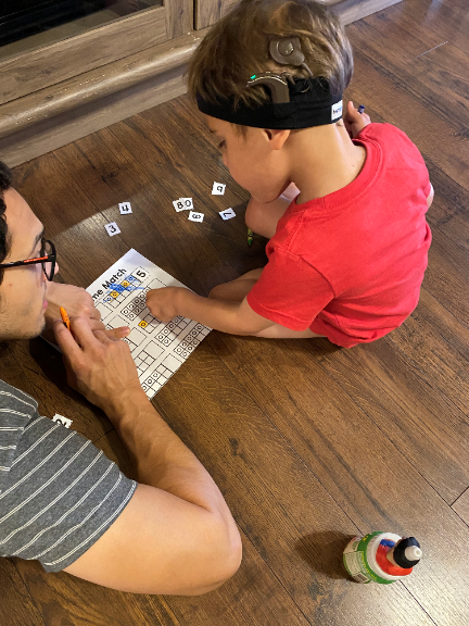 Boy wearing his Nucleus Sound Processor with Dad, his hearing practice partner, playing a number matching game.