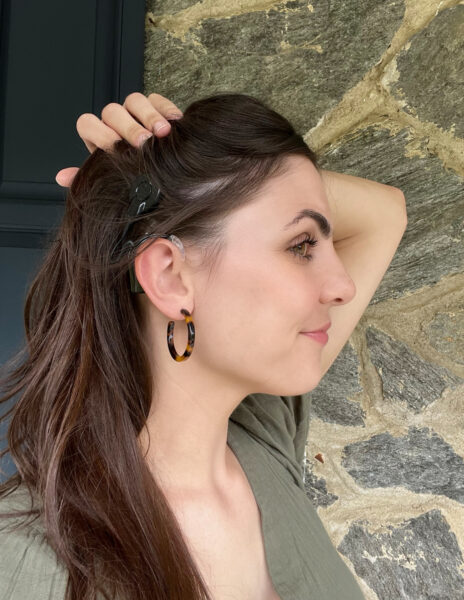 Cassandra, who self-paid for her cochlear implant, shown from a side profile, pulling back her hair to reveal her sound processor on her ear. 