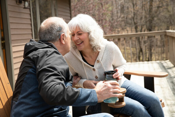 A picture of Scott, a cochlear implant recipient, leaning in to kiss his wife, they are both sitting on patio furniture outside of a cabin. 