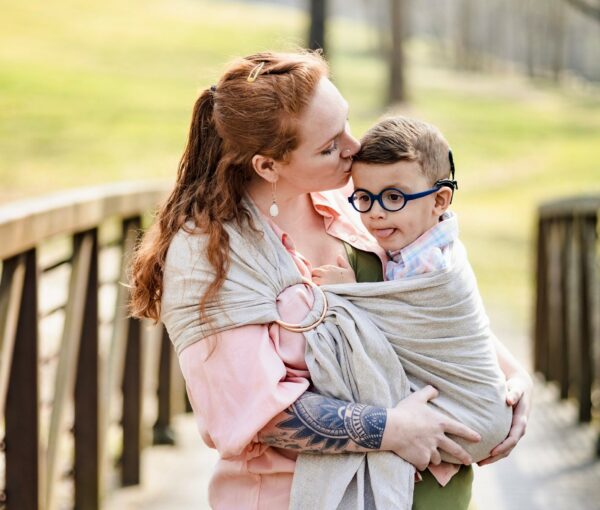 Liam, a young boy with cochlear implants who used speech therapy to make progress in his life, being held in a shawl by his mom, who is kissing his forehead. 