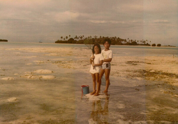 An older photo in sepia, of Amy and her brother standing in a shallow body of water in Indonesia as children. She is holding a fishing net, with buckets at their feet. 