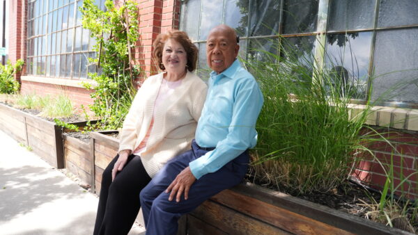 A photo of Dr. G and Roseann, a couple who were filmed in a behind the scenes video shoot to describe how helpful cochlear implants can be for loved ones. They are sitting on on a plantar box near a building with many windows. 