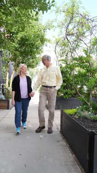 A photo of Jerry and Sherri, a couple who participated in our behind the scenes video shoot showcasing the benefits of cochlear implants. They are holding hands and walking down an aisle of plantar boxes. 