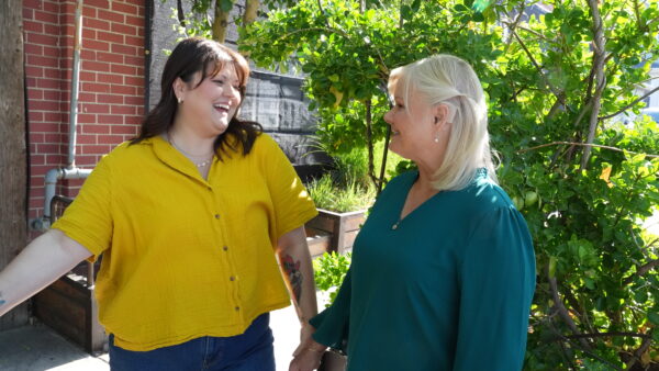 A photo of Cathy and Molly, a mother, daughter duo that were part of our behind the scenes video shoot for cochlear implant loved ones. They are holding hands and laughing together underneath a tree. 