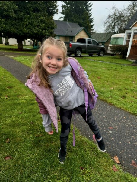 Brielle, a girl with a cochlear implant, smiling big for the picture, outside of a house on a sidewalk. She is leaning to the side in a pose. 