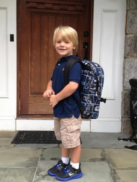 Danny, whose cochlear implants helped him become involved in school, pictured as a young child smiling outside his front door with a backpack. 