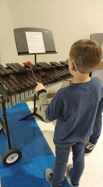 Micah, a boy who has cochlear implants from CMV, pictured from behind showing his sound processor on his ear, and playing the xylophone. 