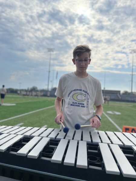 Micah, a boy with cochlear implants from CMV, playing the xylophone outdoors in the sun, while standing on a football field. 