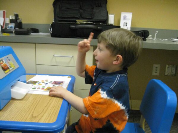 Micah, who received cochlear implants to treat the hearing loss from a CMV diagnosis, pictured as a child doing speech therapy, with a smile and his index finger in the air. 