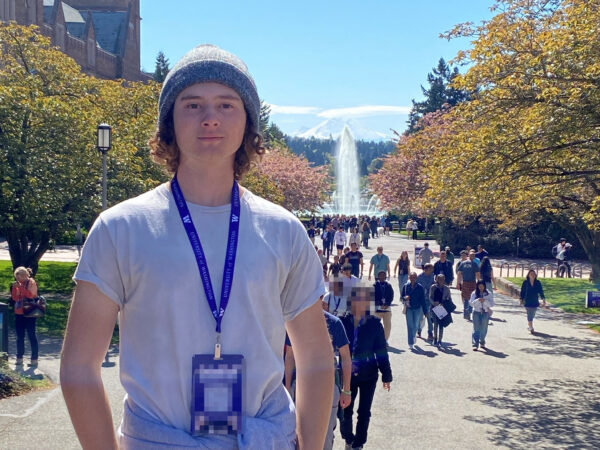 Michael, who is an advocate and part of the Cochlear Mentor program, posing for a photo on his college campus, in front of a row of trees and a very large water feature. 