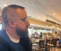 Chet, who experienced hearing loss from a rare autoimmune disease, shown in a cafe with a side profile, where his cochlear implant is visible behind his ear. 