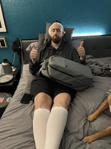 Chet, who received a cochlear implant after a hearing loss diagnosis from a rare autoimmune disease, shown laying on his bed, with a Cochlear backpack across his lap and showing two thumbs up. 