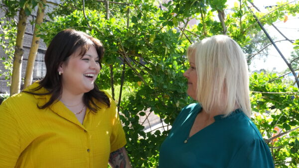 A photo of two loved ones, a older mother and her adult daughter smiling as they converse with each other outdoors under a group of trees. 