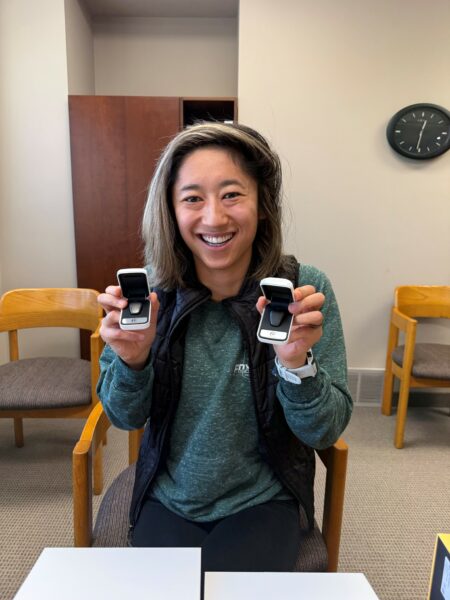 Rachel, smiling and holding her Baha 6 Max Sound Processors at her upgrade activation appointment; Baha Sound Processor features.