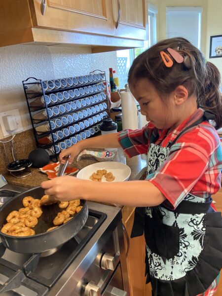 Adrian, a child who uses the Osia System, in her kitchen wearing an apron and taking shrimp off the stovetop.