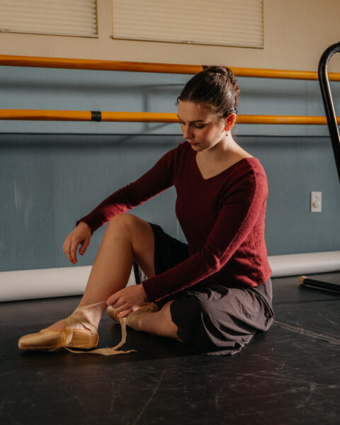 Fiona, a cochlear implant ballerina pictured sitting on the floor in a studio and lacing up her pointe shoes. 