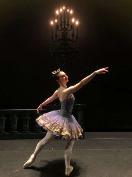 Fiona, a cochlear implant ballerina pictured posing in a costume with a tutu and a candelabra illuminated in the background above her. 