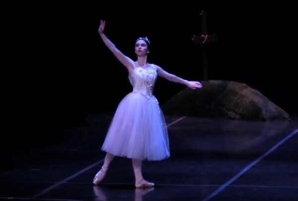 Fiona, a cochlear implant ballerina picture performing on stage, wearing a costume with a tutu and posing with her arm and foot out in a ballet stance. 