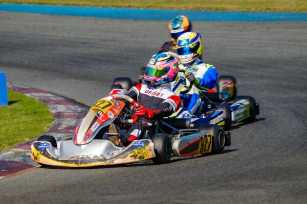 A picture of Helio driving a go kart, wearing a full drivers uniform and helmet. He is coming around a corner with two other drivers behind him. 