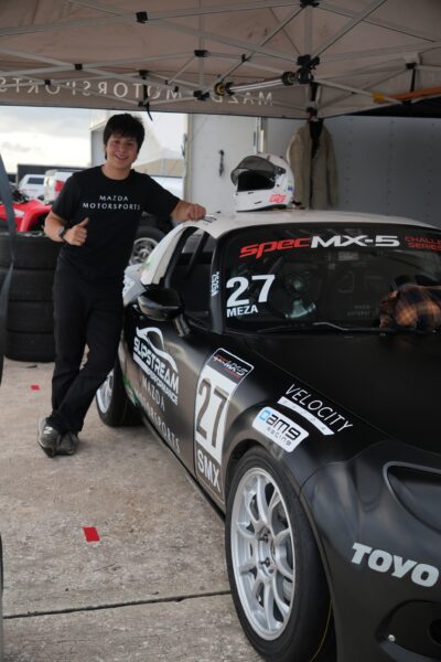 Helio smiling and holding a thumbs up as he leans against his Mazda Miata race car. 