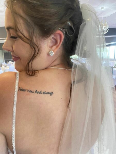 Kristina, whose cochlear implants helped give her access to sound, looking over her shoulder and smiling in her wedding dress. A "Hear now. And always" tattoo is visible on her back shoulder. 