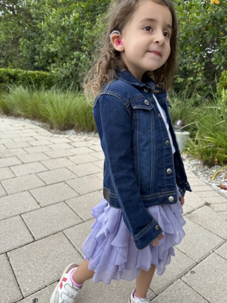 Fiona, who along with her sibling have hearing loss from MYO15A, shown smiling posing for a photo on a sidewalk, with her cochlear implant sound processor visible on her ear.