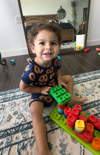 Augustine who has cochlear implants to help with Mitchell Syndrome sitting on a floor rug, playing with toy blocks and smiling up at the camera.