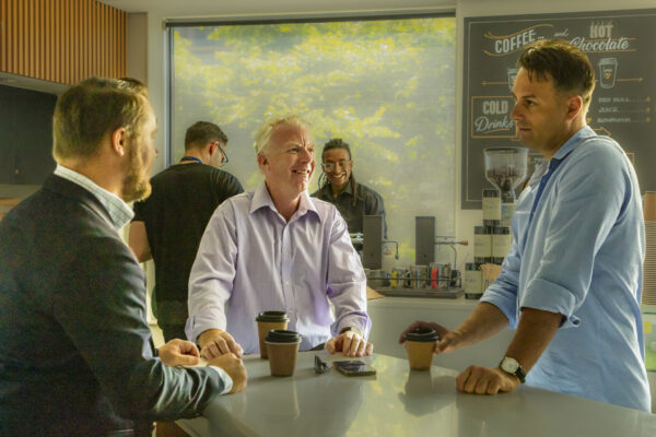 Three men stand talking in a coffee shop; navigating listening fatigue.