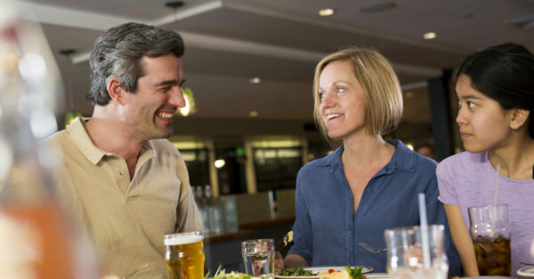 A family sits smiling in a restaurant having a meal while talking. A Cochlear Nucleus 8 Sound Processor is barely visible above the father’s ear; new sound processor.