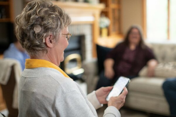A woman sits in a cozy room looking at her mobile phone. Family members sit on a sofa opposite the woman. A Cochlear Nucleus 8 Sound Processor is visible beneath her hair.
