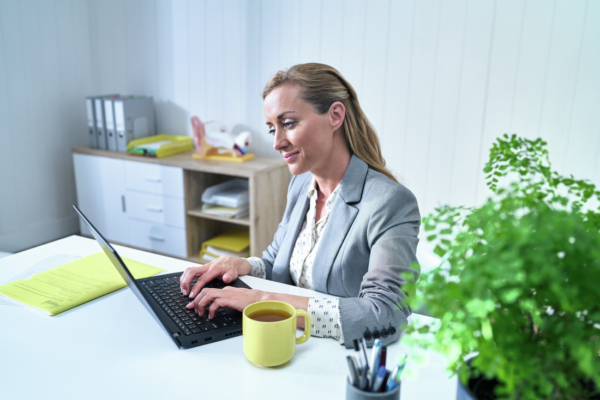 Woman, returning to work after a cochlear implant, sits in her office typing on her laptop. A yellow mug of tea sits on her desk.