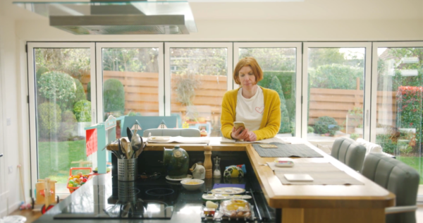 Woman in a yellow cardigan sitting at kitchen counter looking at her phone; hearing with the Nucleus Kanso 3 Nexa Sound Processor
