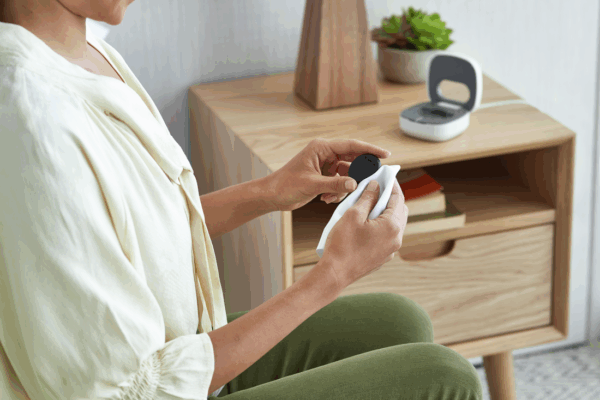 A woman is holding a Cochlear™ Kanso® 3 Sound Processor with a home charger on her bedside table; tips to care for your cochlear implant sound processor.