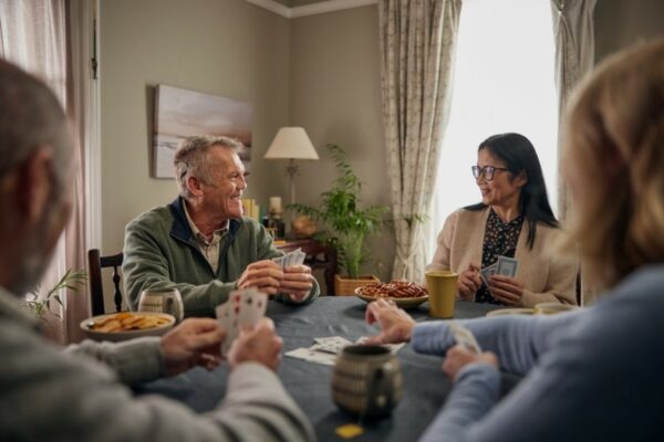 A picture of an older gentleman playing cards with his 3 friends, around a table filled with snacks and drinks. He is wearing a cochlear implant on his ear. 