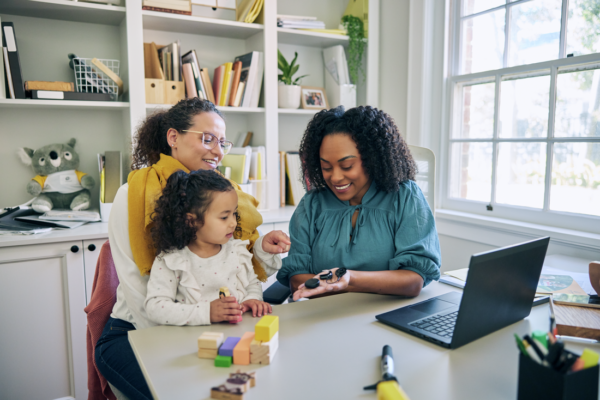 A young girl and her mother meet with a healthcare professional in a bright, welcoming office. The mother, smiles as she watches the clinician interacts with her child. In her hand the clinician has several cochlear implant sound processors.
