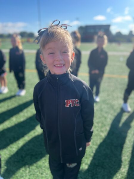 Lyla, a young girl with unilateral hearing loss who received a cochlear implant, smiling on a sunny day in front of a team of cheerleaders.