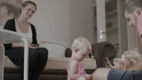 A young girl with a cochlear implant plays with dolls on the floor. A woman and a man watch her in a bright, neutral living room.